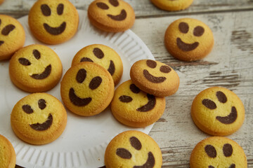 Yellow cookies shaped like smiling emojis are scattered on a wooden table from a white plate in close-up, good mood, festive atmosphere, friendliness and warmth, happiness and joy.