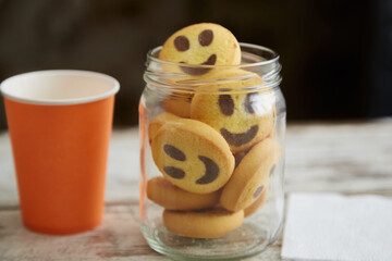 Cookies shaped like smiling emojis in a glass jar on a table with a cup and napkin nearby in close-up, festive homemade treat, family traditions, family holidays and parties.