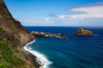 Powerful waves crashing against Madeira’s rocky shoreline, with steep green cliffs and the vast Atlantic Ocean under a partly cloudy sky.