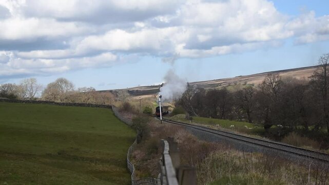 Goathland, North Yorkshire, United Kingdom - May, 6, 2021 : Scenic Steam Engine Train coming through picturesque countryside, fields, farm and forest, blue sky white cloud.