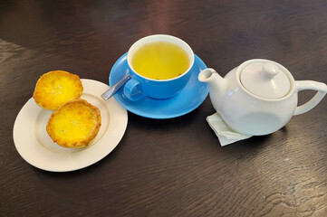 A white teapot, a blue cup with green tea on a saucer, baked baskets on a white plate.