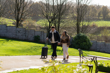 Young couple strolling together in the gardens of a hotel resort, with the man pulling a rolling...
