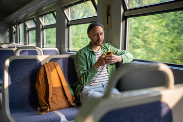 Focused man leans into smartphone, scrolling social media and chatting, checking train schedule on commute. Male online connection, public transit, morning travel, schedule tracking on road trip
