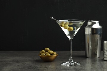 Martini cocktail with olives in glass and shaker on grey table against black background, space for text