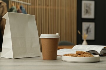 Paper cup, plate of cookies, book and takeaway bag on light wooden table in cafe. Mockup for design