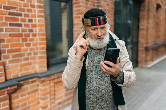 Portrait of surprised homeless elderly man having mobile phone conversation and raising index finger while standing in urban street in front of brick wall. Concept of raw reality of poverty, despair.