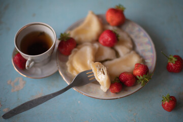 A plate of sweet dumplings with strawberries on a light blue table, with a bite taken out to reveal the creamy filling. A cup of tea sits nearby, completing the relaxing scene.
