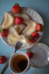 A plate of sweet dumplings with strawberries on a light blue table, with a bite taken out to reveal the creamy filling. A cup of tea sits nearby, completing the relaxing scene.
