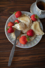 A plate of dumplings with strawberries served on a wooden table, dusted with powdered sugar. A cup of tea in a vintage porcelain cup adds a touch of elegance to this homemade meal.
