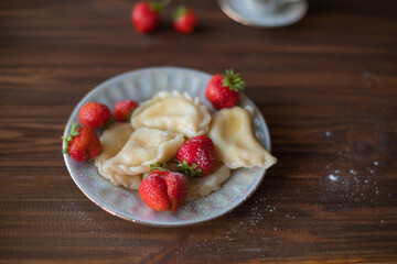 A plate of dumplings with strawberries served on a wooden table, dusted with powdered sugar. A cup of tea in a vintage porcelain cup adds a touch of elegance to this homemade meal.
