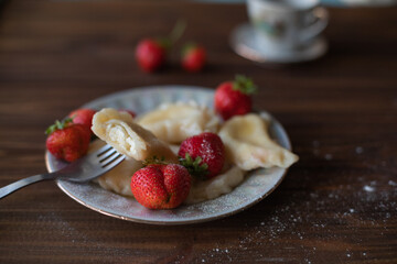 A plate of dumplings with strawberries served on a wooden table, dusted with powdered sugar. A cup of tea in a vintage porcelain cup adds a touch of elegance to this homemade meal.
