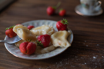 A close-up of a plate of homemade dumplings with fresh strawberries, lightly dusted with powdered sugar, placed on a dark wooden table. The warm tones create an inviting and cozy atmosphere.