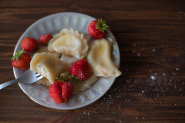 A close-up of a plate of homemade dumplings with fresh strawberries, lightly dusted with powdered sugar, placed on a dark wooden table. The warm tones create an inviting and cozy atmosphere.