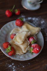 A close-up of a plate of homemade dumplings with fresh strawberries, lightly dusted with powdered sugar, placed on a dark wooden table. The warm tones create an inviting and cozy atmosphere.