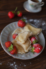 A close-up of a plate of homemade dumplings with fresh strawberries, lightly dusted with powdered sugar, placed on a dark wooden table. The warm tones create an inviting and cozy atmosphere.
