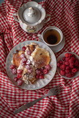 Golden-brown cottage cheese pancakes served with fresh raspberries, lightly dusted with powdered sugar. The plate is accompanied by a vintage teapot, a cup of tea, and a red gingham cloth, creating a 