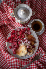 Golden-brown cottage cheese pancakes served with fresh raspberries, lightly dusted with powdered sugar. The plate is accompanied by a vintage teapot, a cup of tea, and a red gingham cloth, creating a 