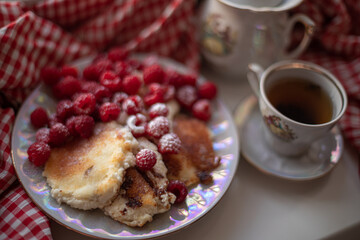 Golden-brown cottage cheese pancakes served with fresh raspberries, lightly dusted with powdered sugar. The plate is accompanied by a vintage teapot, a cup of tea, and a red gingham cloth, creating a 