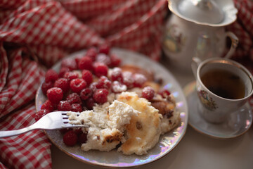 Golden-brown cottage cheese pancakes served with fresh raspberries, lightly dusted with powdered sugar. The plate is accompanied by a vintage teapot, a cup of tea, and a red gingham cloth, creating a 