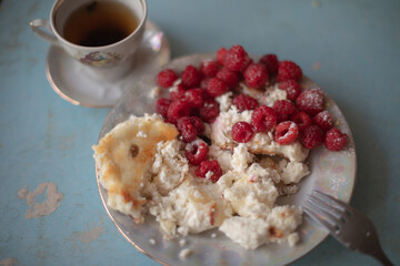 Golden-brown cottage cheese pancakes served with fresh raspberries, lightly dusted with powdered sugar. The plate is accompanied by a vintage teapot, a cup of tea, and a red gingham cloth, creating a 