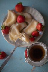 A plate of sweet dumplings with strawberries on a light blue table, with a bite taken out to reveal the creamy filling. A cup of tea sits nearby, completing the relaxing scene.
