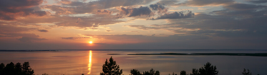 Fototapeta premium A delightful summer sunset over a full-flowing plain river. The sun in a cloudy sky descends to the horizon. The sunlight is reflected in gold on the water surface. Copy space.