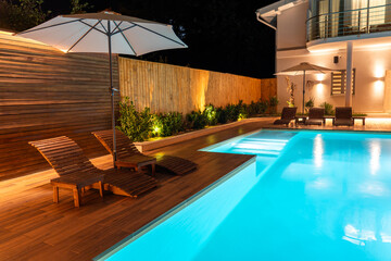 Modern swimming pool with wooden deck, loungers, and soft lighting at night. Concept of elegant outdoor living