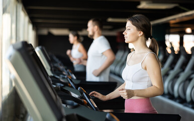 Fototapeta premium During group workout, girl walks on surface of treadmill, creates cardio load. Gym visitor uses treadmill simulator for cardio training. Girl exercising in health club, perform sport routine