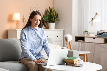 Beautiful happy female writer with laptop and books sitting on sofa at home