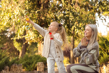 Little girl with her mother blowing soap bubbles in park on autumn day