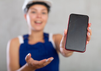 Cheerful female worker in overalls and hardhat showing screen of her phone