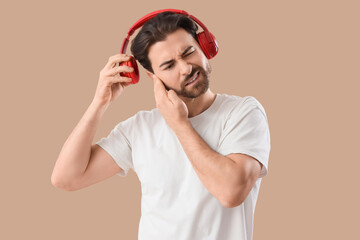 Young man with headphones suffering from ear pain on beige background