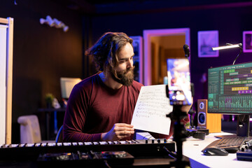 Young singer pointing at music sheet to explain notes on synthesizer, teaching people how to play piano electronic keyboard in home studio. Artist performer playing midi controller keys, online vlog.