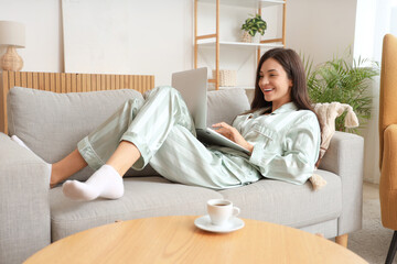 Young woman in green pajamas working with laptop on sofa at home