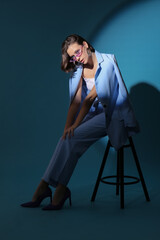 Beautiful young woman in stylish outfit sitting on stool against blue background