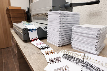 Modern binding machine, stacks of notebooks and double loop wire binding spines on wooden table...