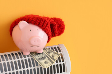 Piggy bank with warm hat and money on convector heater against orange background. Heating saving...