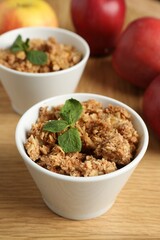 Delicious apple crisp with mint in bowls and fresh fruits on wooden table, closeup
