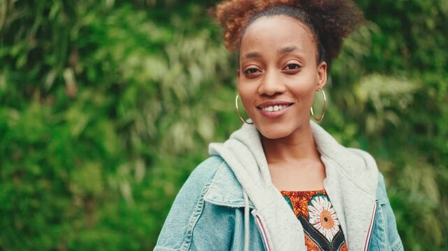 Closeup portrait African girl with ponytail wearing denim jacket, in crop top with national pattern turns her head and smiles at the camera in city park on green trees background. Slow motion
