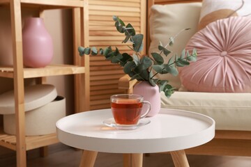 Coffee table with tea and eucalyptus near armchair at home