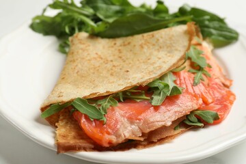 Delicious crepes with salmon and arugula on white table, closeup