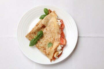 Delicious crepe with salmon, cream cheese and basil on white tiled table, top view