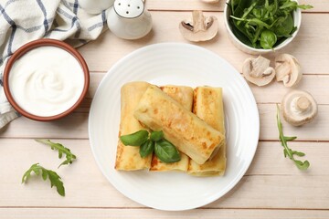 Delicious rolled crepes with basil, arugula, mushrooms and sour cream on wooden table, flat lay