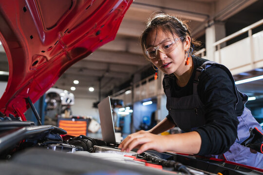 Female chinese mechanic using laptop while repairing car engine in garage