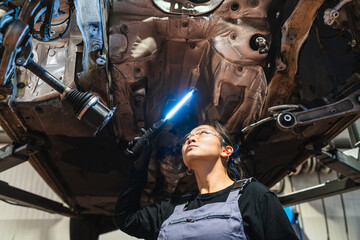 Female chinese mechanic inspecting car undercarriage with light in repair shop