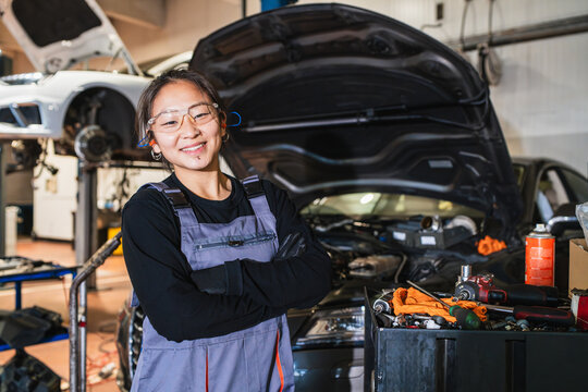 Confident chinese female mechanic smiling with crossed arms in auto repair shop
