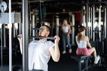 Fototapeta premium Young athletic man in sportswear training his arms muscles on machine in gym