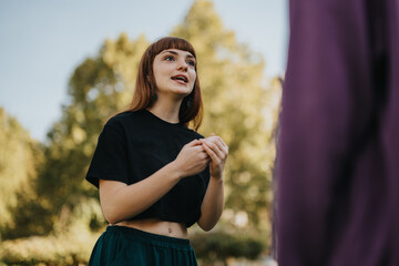 A young woman wearing a black top speaking with someone outdoors. She is in a park setting with green trees in the background.