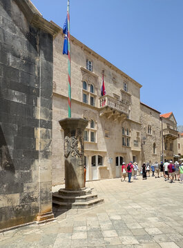 Korcula, Croatia - June 30, 2024: Kolumna or historical Column with flag on Saint Marks Square, in front of his Cathedral. Street scene with historic facade and tourists walking along Palaca Arneri
