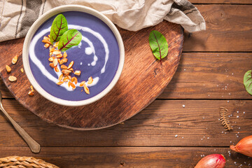 A bowl of vibrant purple sweet popato soup garnished with a swirl of cream, toasted almonds, and fresh herbs, resting on a rustic wooden board.